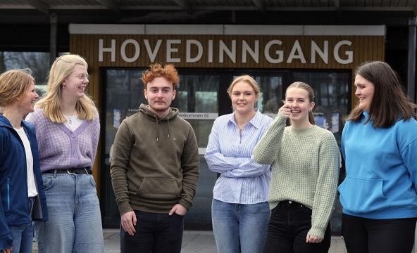 Seks studenter og ansatte på rekke foran inngangen på et bygg. Foto