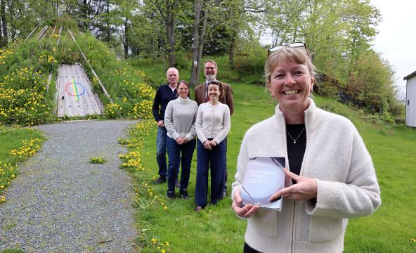 Kvinne som holder opp en bok med fire kollegaer i bakgrunnen. Alle sammen foran en samisk gamme. Foto