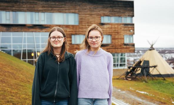 To unge jenter står foran en bygning og ser inn i kamera.