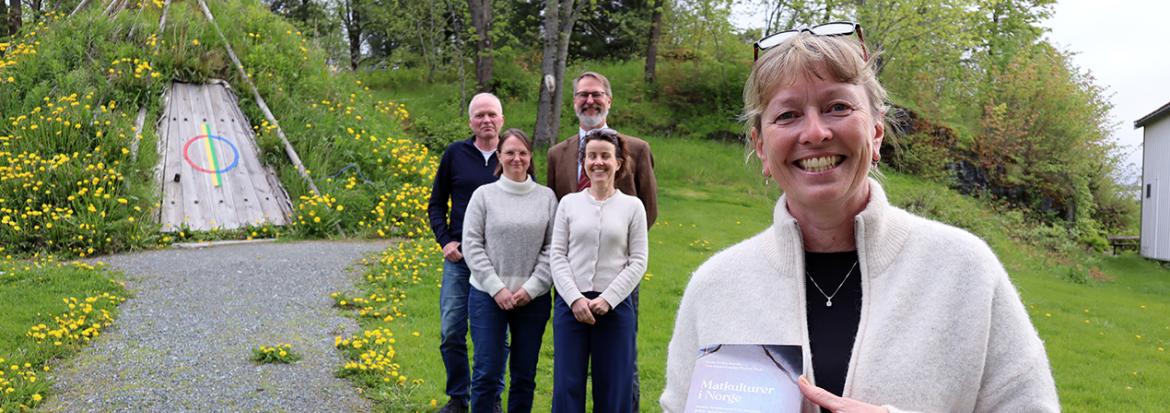 Kvinne som holder opp en bok med fire kollegaer i bakgrunnen. Alle sammen foran en samisk gamme. Foto