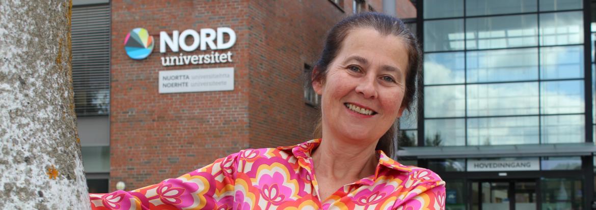 Blid dame i rosa bluse foran hovedinngangen til Nord universitet.