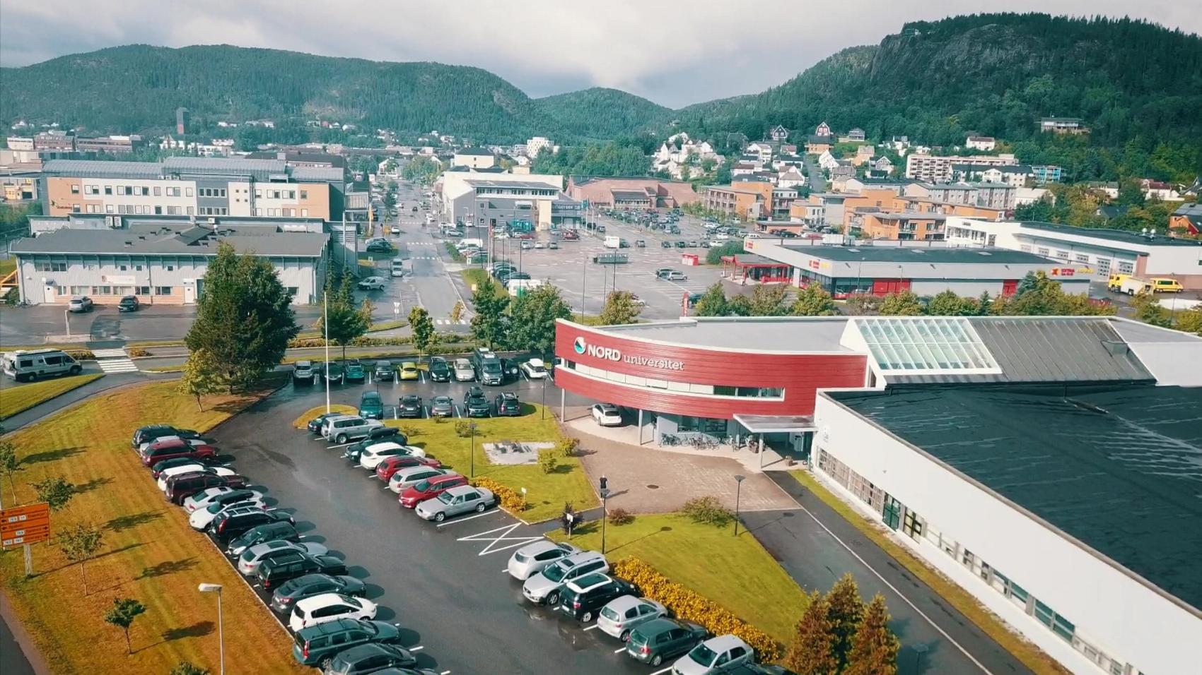 Hus, bygg, biler og parkeringsplass. Oversiktsbilde tatt fra luften.