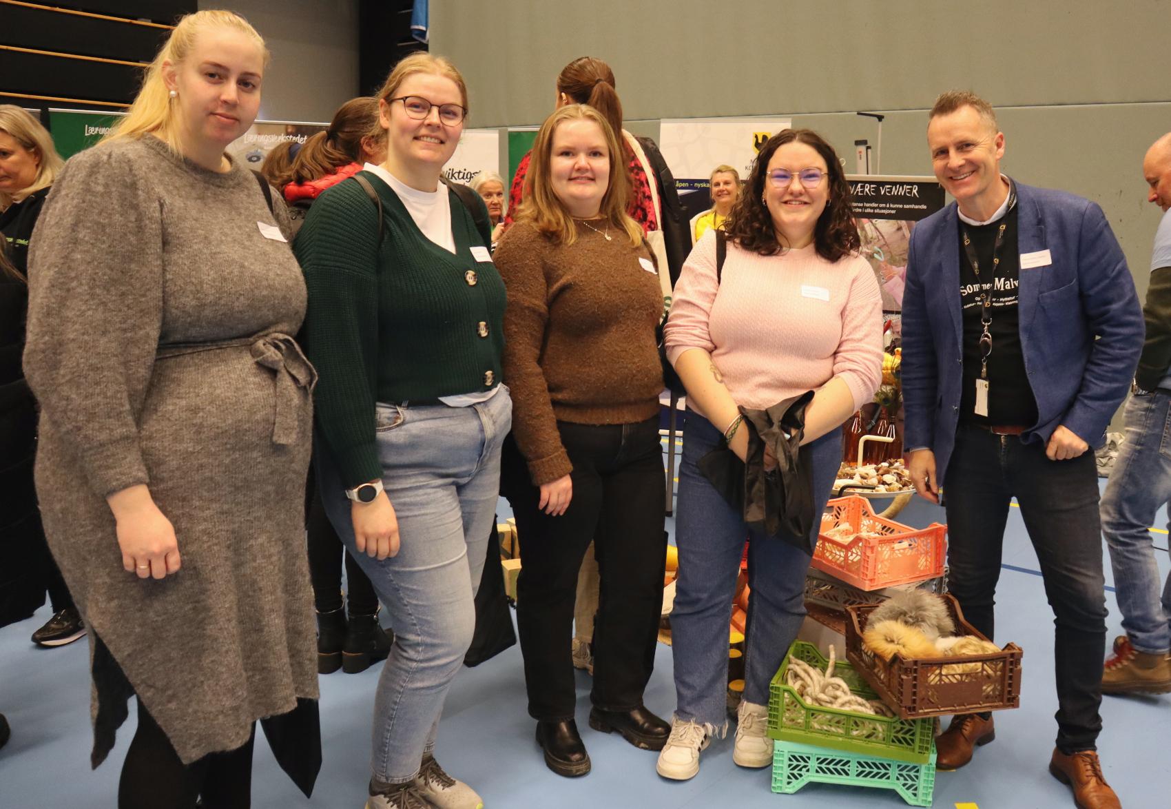 Fire kvinner og en mann smiler mot kamera. Mennesker og rollups bak dem. 