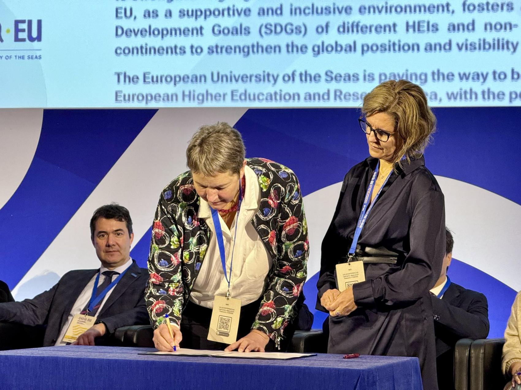 Dame signerer et papir på et bord, en annen dame står ved siden av. En mann sitter i bakgrunnen. 