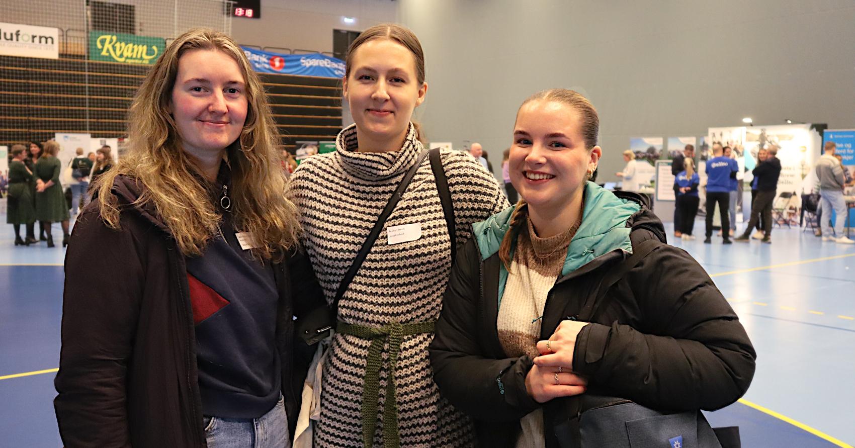 Tre smilende unge kvinner står i en idrettshall. I bakgrunnen skimtes informasjonsstands med beslkende rundt.