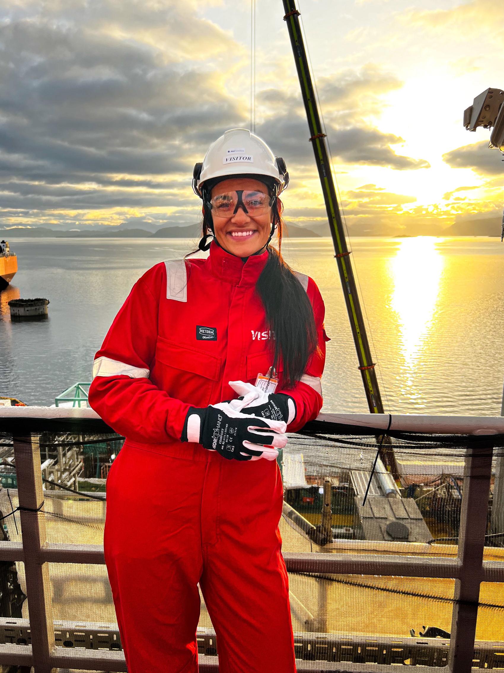 Kvinne med hjelm, vernebriller og kjeledress med fjord og sol i bakgrunn. Foto