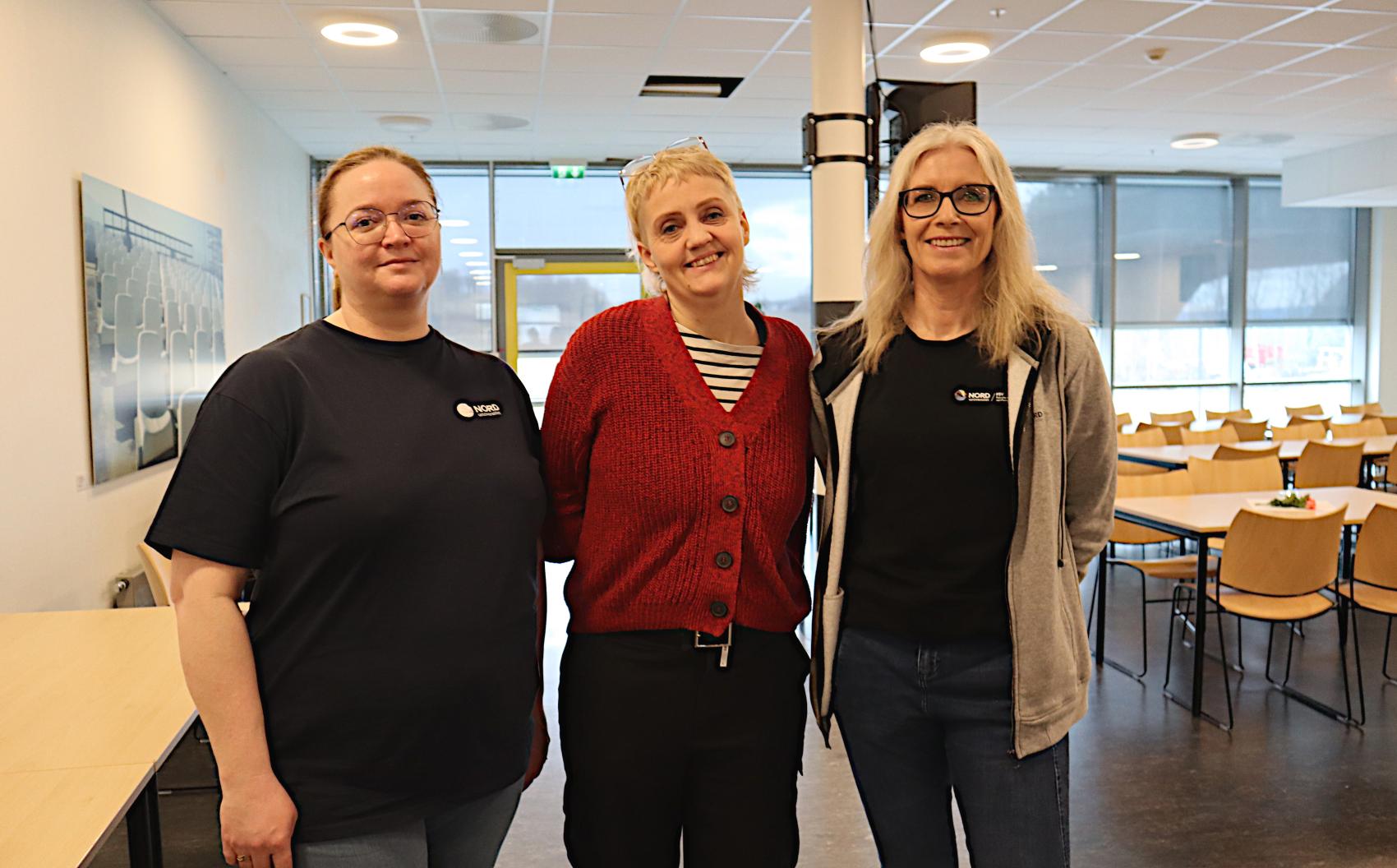 Tre kvinner smiler til kamera, de står i en sal med stoler og bord i bakgrunnen