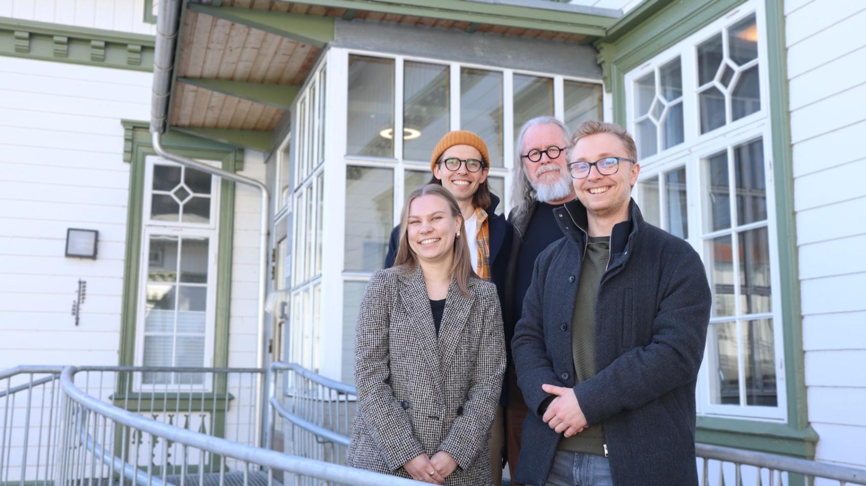 Fire smilende personer står foran et hvitt bygg med gammeldagse vinduer. Tre menn og en kvinne. 