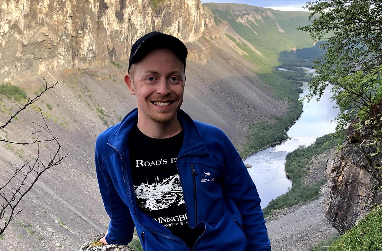 Mann med blå skjorte og caps smiler foran landskap