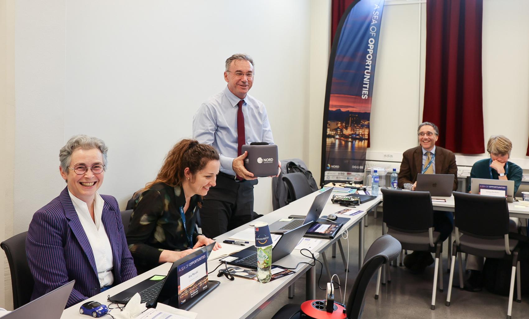 5 people in a meeting room, sitting behind desks, smiling. One man is standing and holding a microphone cube