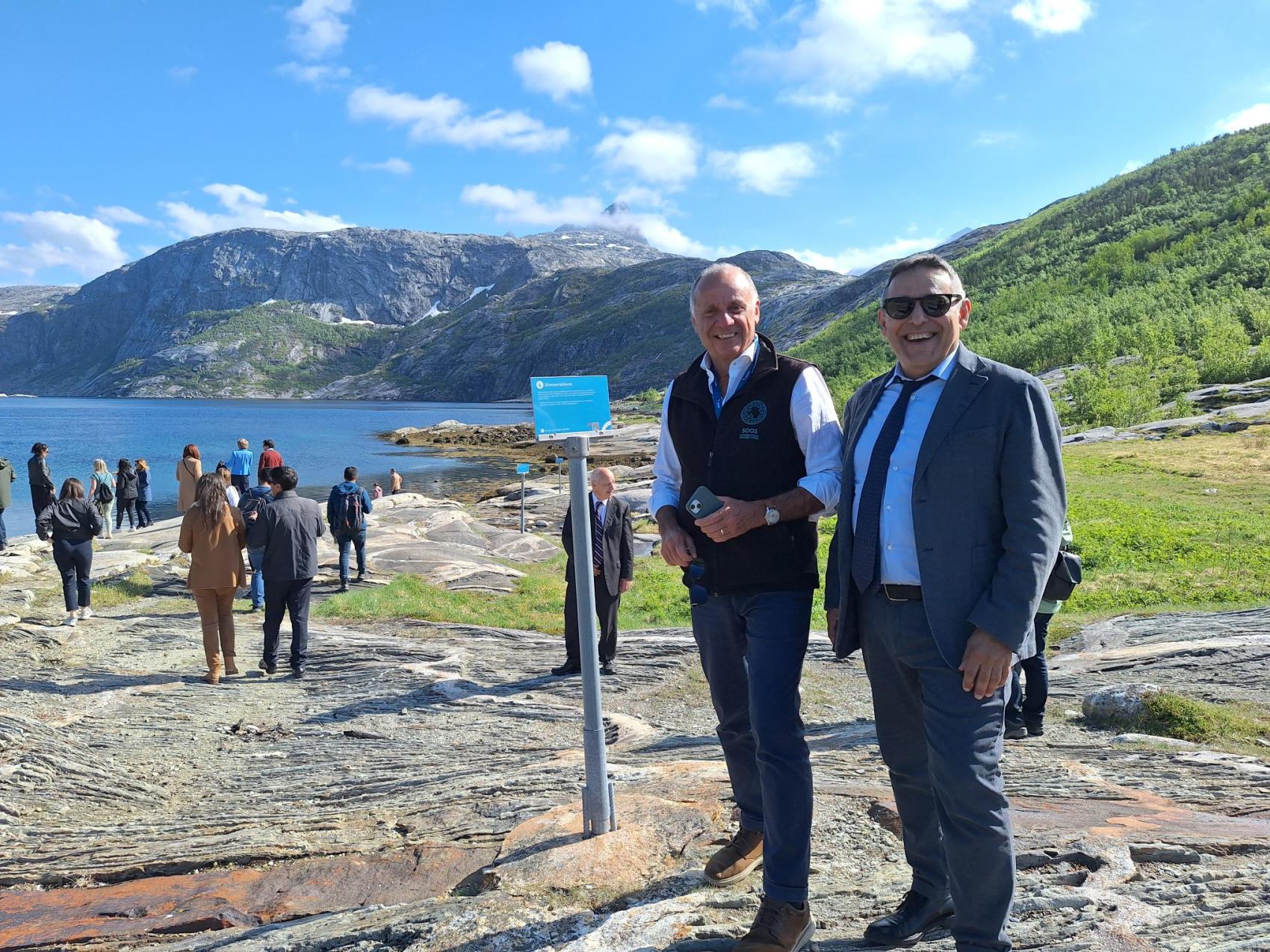 Two formally dressed men standing on rocks in a sunny weather, the sea and more people in the background