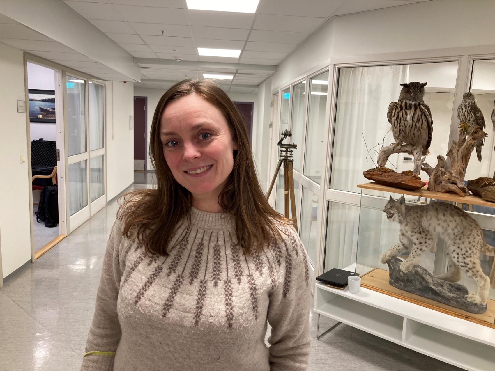 Kvinne står i en korridor, bak står en monter med utstoppede fugler og kattedyr