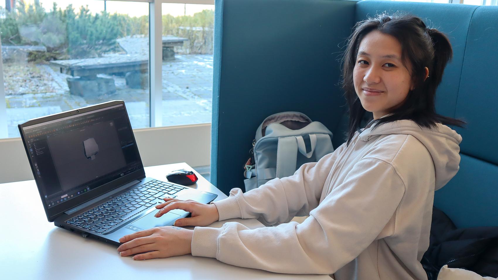 Ung kvinne i hvit genser sitter med en laptop foran seg og ser mot kamera.