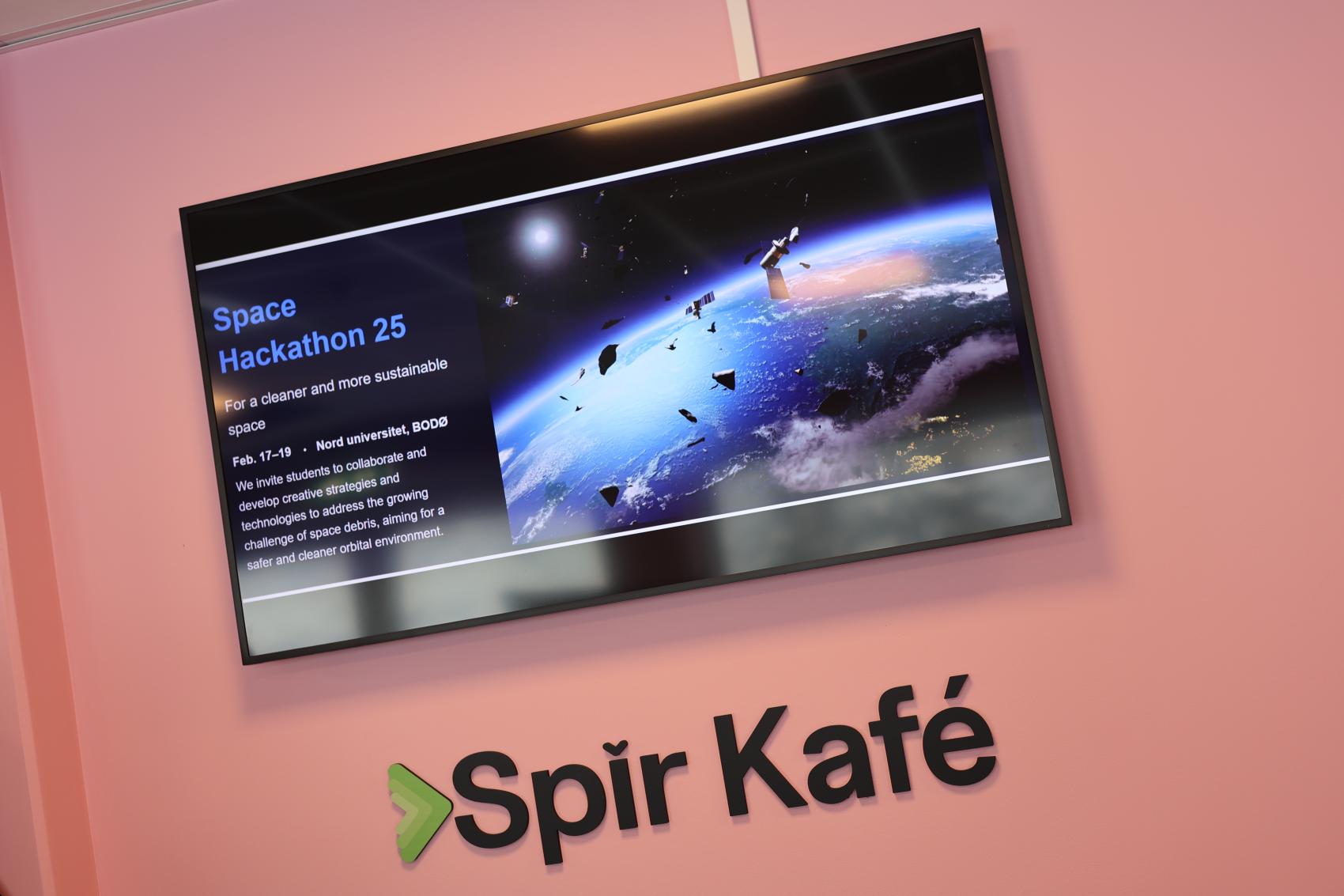 bilde av en stor infoskjerm på rosa vegg, med teksten Spir Kafé.