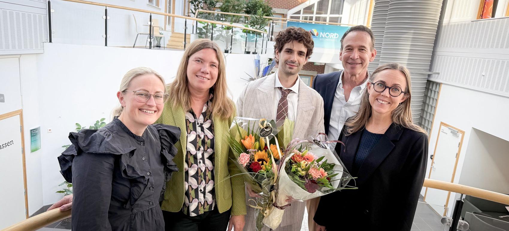 Fire personer som omkranser en ung forsker med flere blomsterbuketter i hendene. Roberto Vivona disputas.