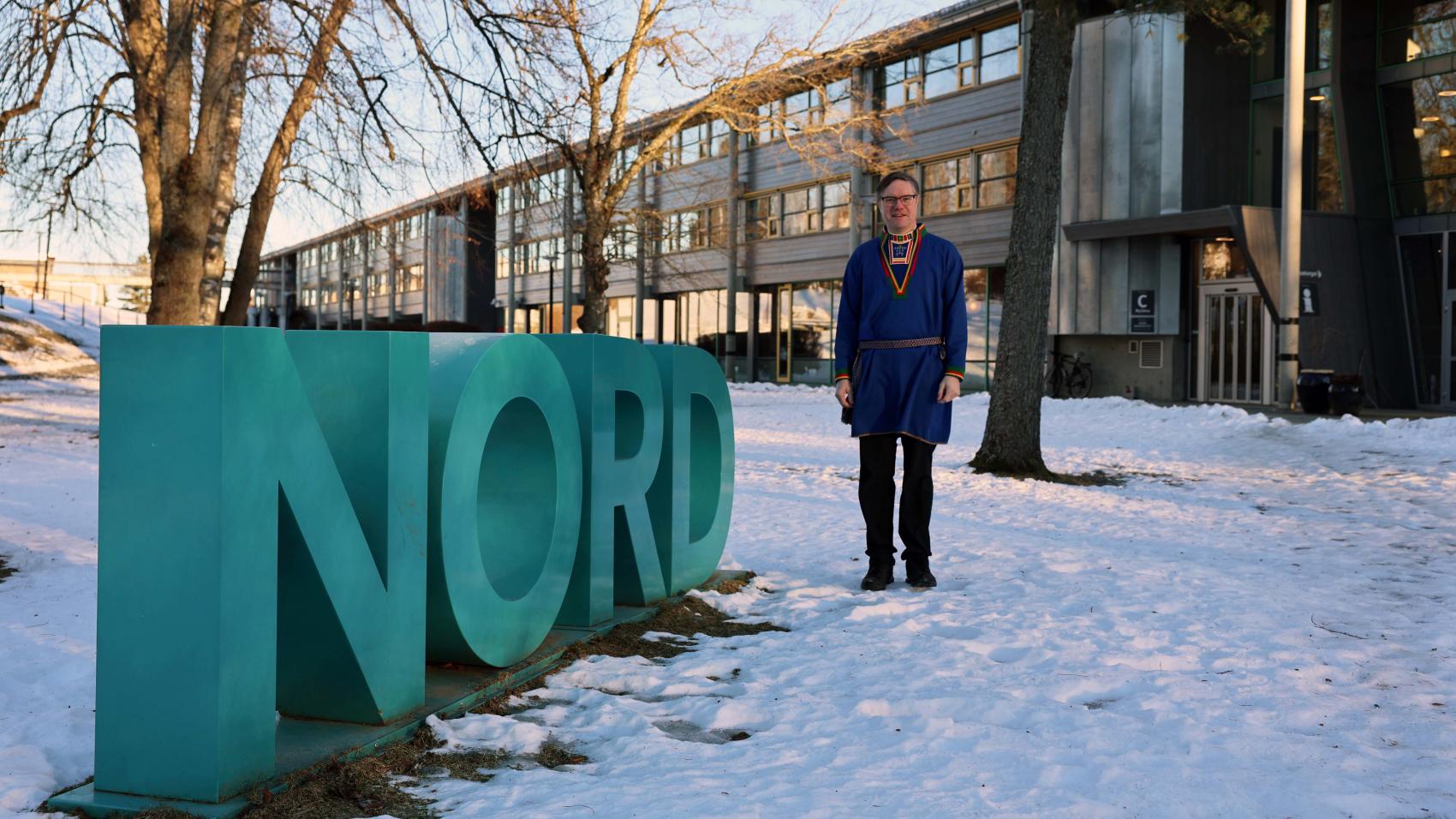 Mann i samisk kofte ute i en park foran et universitetsbygg ved siden av fire store bokstaver NORD. Foto