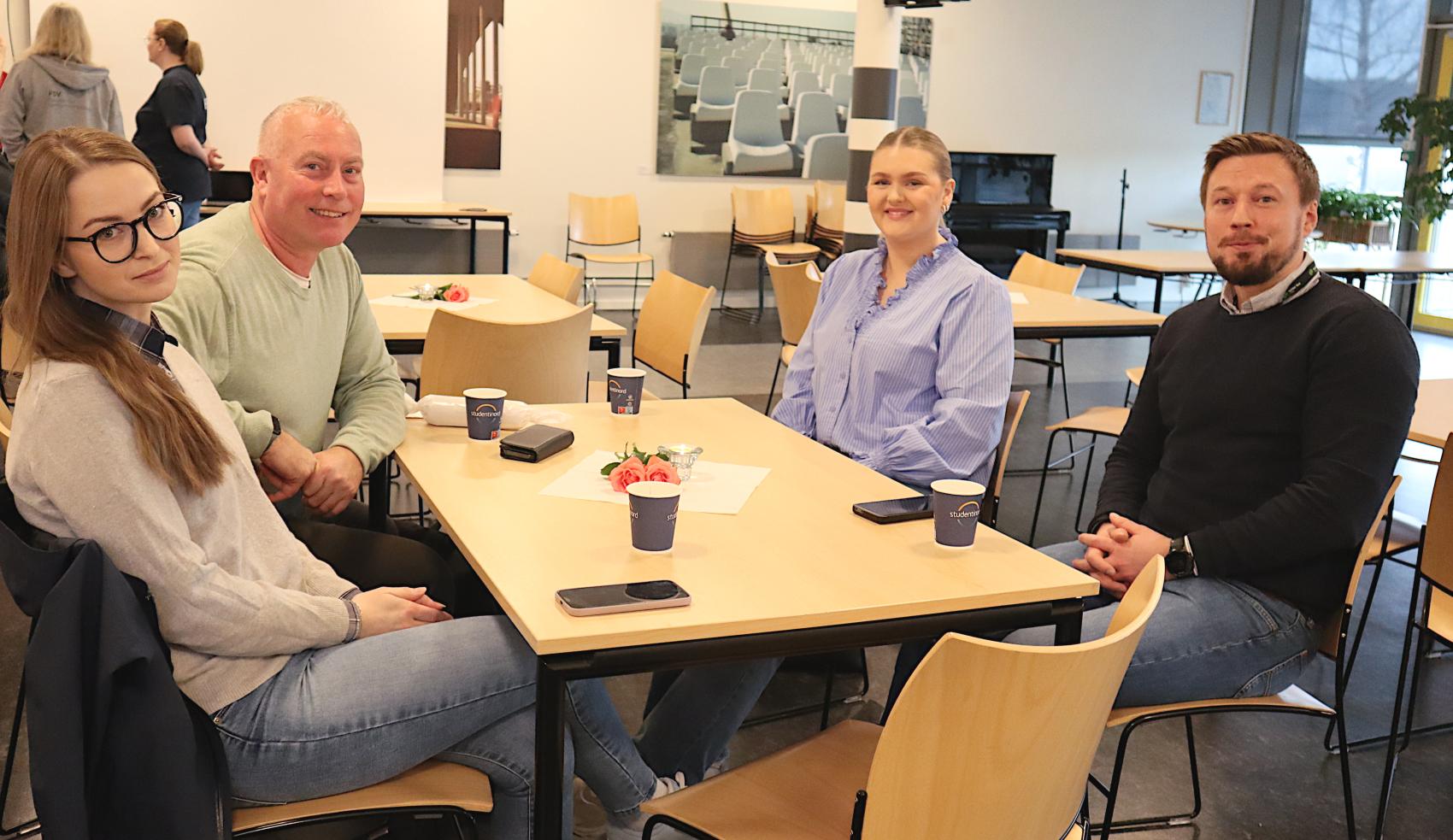 Fire mennesker sitter rundt et bord i en kantine mens de smiler til kamera. Kaffekopper, mobiler og en blomst på bordet. 