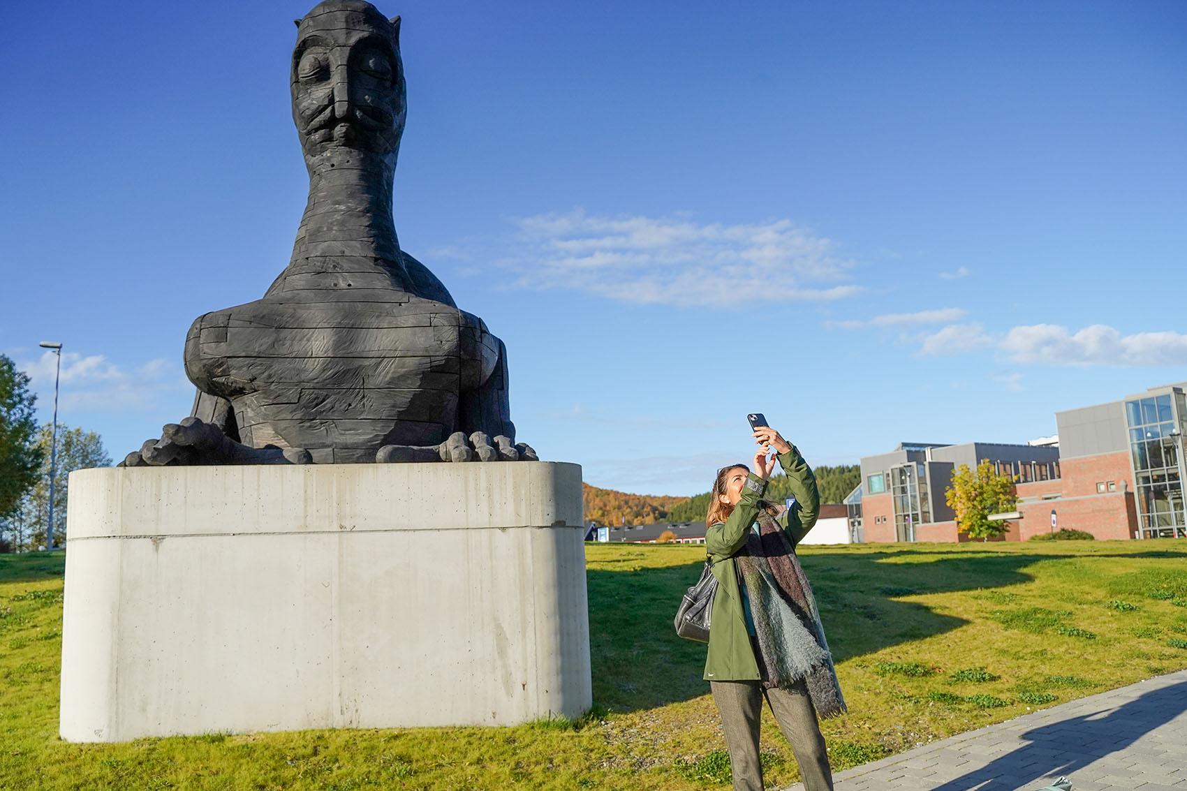 Kvinne som tar selfie ved en stor skulptur på campus.