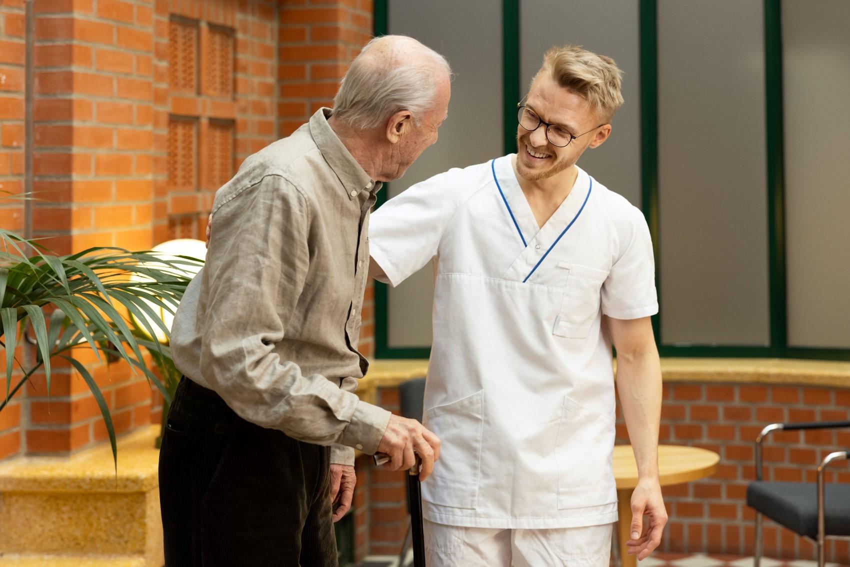 Mannlig sykepleier smiler mot eldre mann med stokk