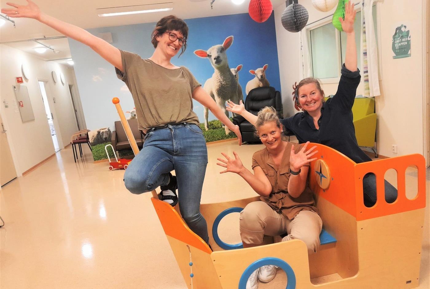 Tre smilende damer med løftede hender sitter i et lekeapparat inenndørs. På veggen bak er det bilde av sauer på tapetet. 