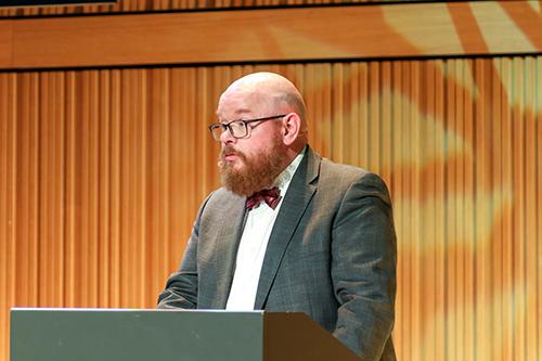 Voksen mann med mye skjegg i dress som står på et podium og snakker til en forsamling.