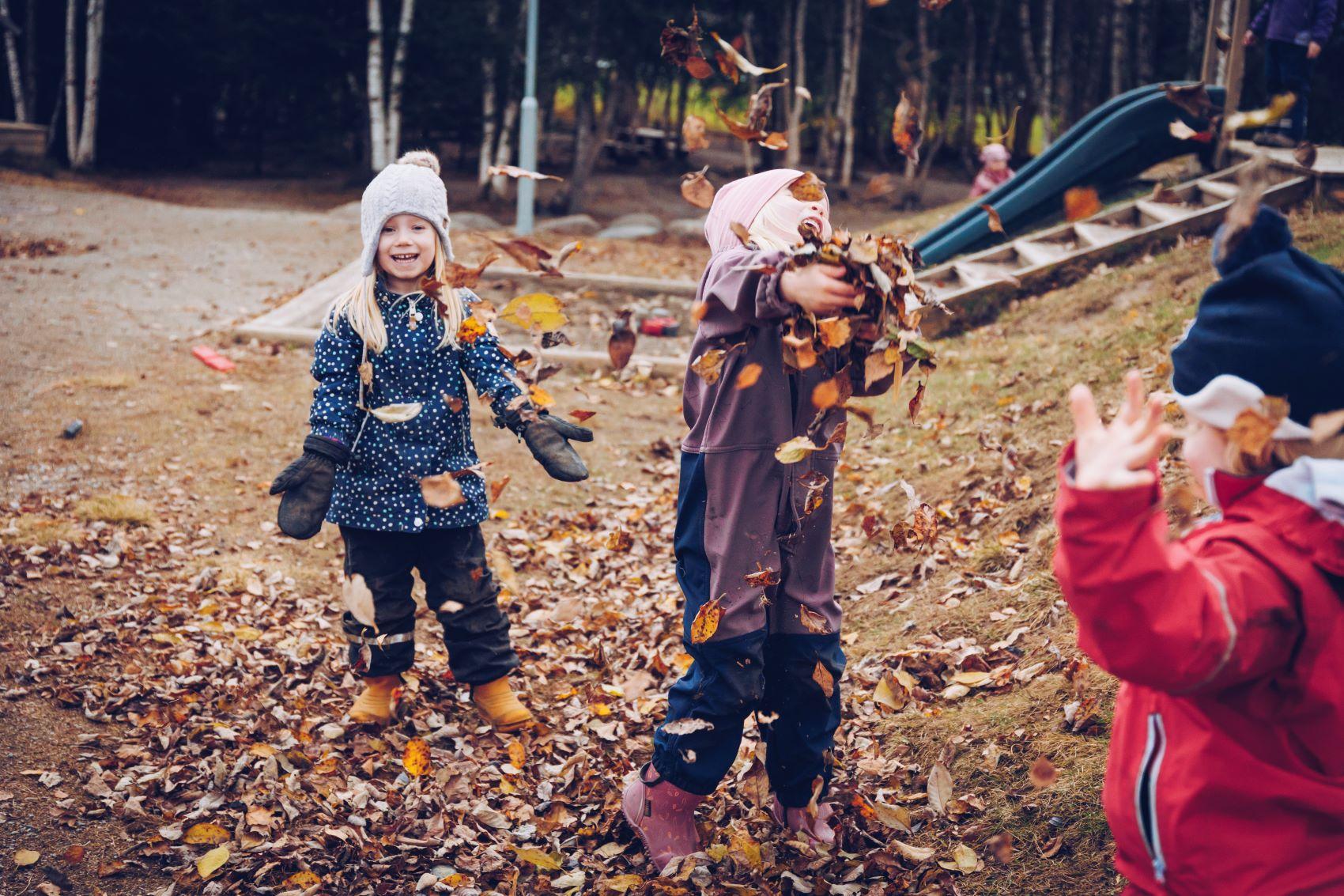 Tre barn kaster høstløv i lufta, sklie og sandkasse i bakgrunnen